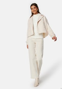 Světlé béžové zkrácené sako, dvouřadý design, spárované s objemným bílým pleteným svetrem a krémovými kalhotami s vysokým pasem.