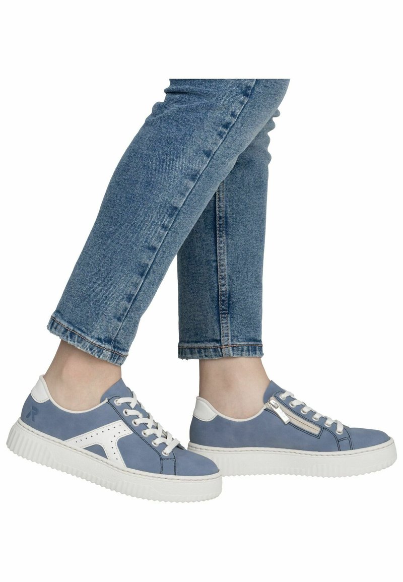 Personne portant des baskets décontractées bleues et blanches avec lacets et fermetures éclair latérales, assorties à un jean en denim bleu.