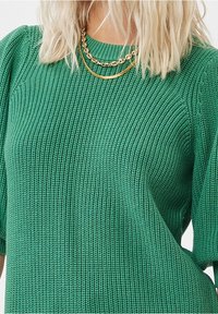 Personne blonde portant un pull vert texturé en tricot avec deux colliers en chaîne dorée superposés sur un fond blanc.