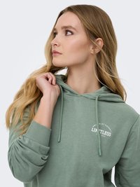 Jeune femme aux longs cheveux blonds portant un sweat à capuche vert avec le texte "LIMITLESS", regardant vers sa gauche tout en tenant le cordon de la capuche.