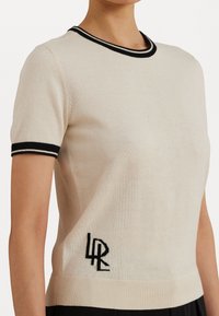 Krátky rukáv pletený sveter v béžovej farbe, so čiernym rebrovaným golierom a manžetami, s čiernym vyšitým logom na ľavej strane.