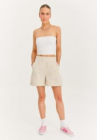 Žena v bielom topi bez ramienok, béžových pruhovaných šortkách, bielych ponožkách a ružových teniskách Adidas, stojaca s rukami v vreckách.