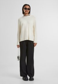 Off-white ribbad tröja med långa ärmar, kombinerad med lösa svarta byxor och robusta svarta skor. Accessoar inkluderar en genomskinlig handväska.