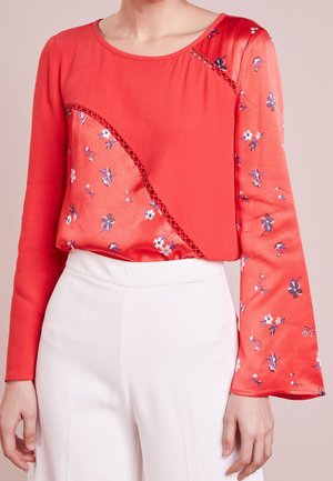 Mujer con camiseta roja de manga larga con estampados florales y detalles recortados combinada con pantalones blancos de talle alto.