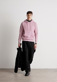 Roosa kapuutsiga särk, millel on ees tasku, koos mustade vaba lõikega pükste ja mustade kingadega, hoidis ühes käes musta jope.