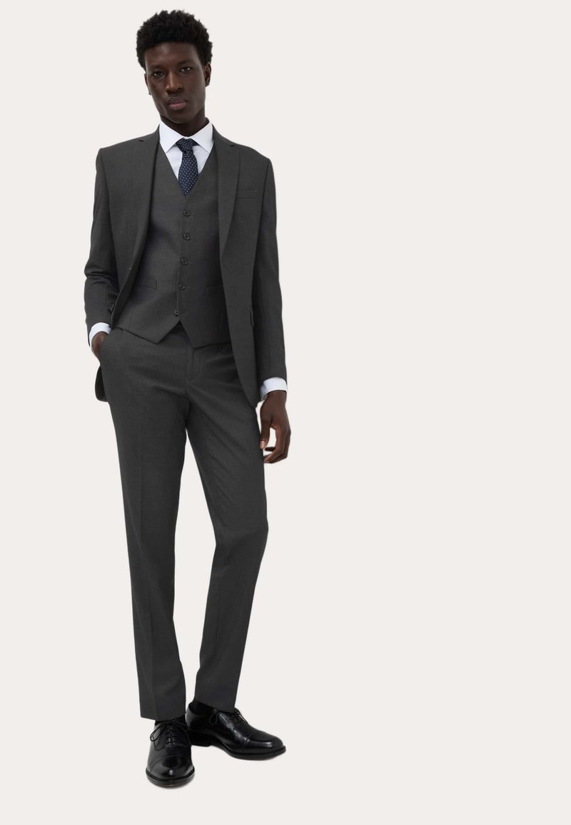 Grå tre-delt jakkesæt med en tætsiddende pasform, tekstureret vest og matchende bukser. Hvid skjorte og blå prikket slips. Polerede sorte sko.