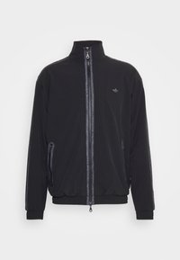 Veste de survêtement noire avec col haut, fermeture éclair, poches latérales zippées et accents réfléchissants le long des manches. Texture de tissu lisse.