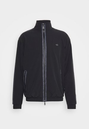 Veste de survêtement noire avec col haut, fermeture éclair, poches latérales zippées et accents réfléchissants le long des manches. Texture de tissu lisse.