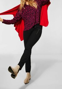 Top con stampa leopardata viola e nera, abbinato a pantaloni neri aderenti con tasca con zip. La modella indossa stivali beige con tacco alto, braccia distese.