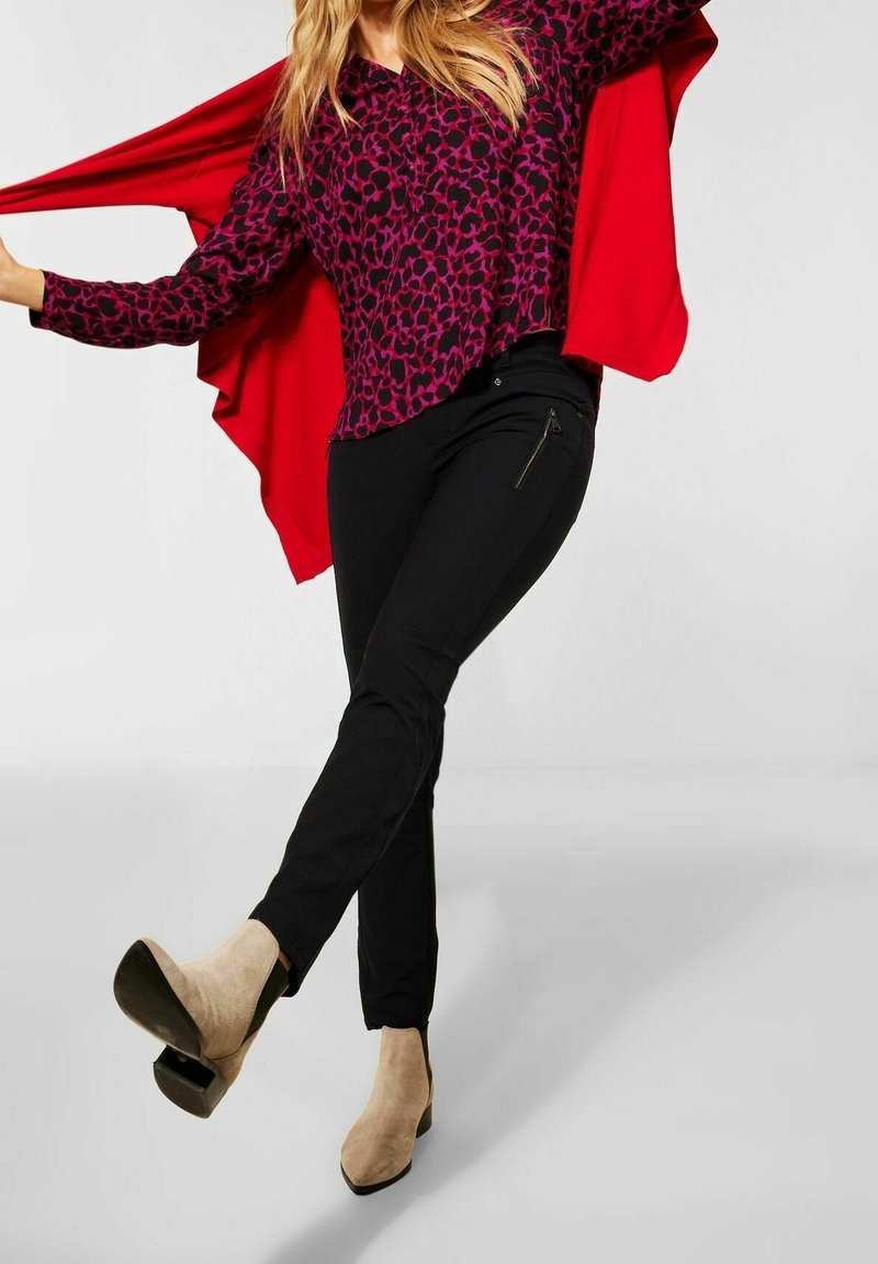 Top con stampa leopardata viola e nera, abbinato a pantaloni neri aderenti con tasca con zip. La modella indossa stivali beige con tacco alto, braccia distese.