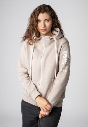 Hellbeige Zip-Hoodie mit Kordelzugkapuze, weichem Stoff, Fronttaschen und weißem Drucktext auf dem linken Ärmel.