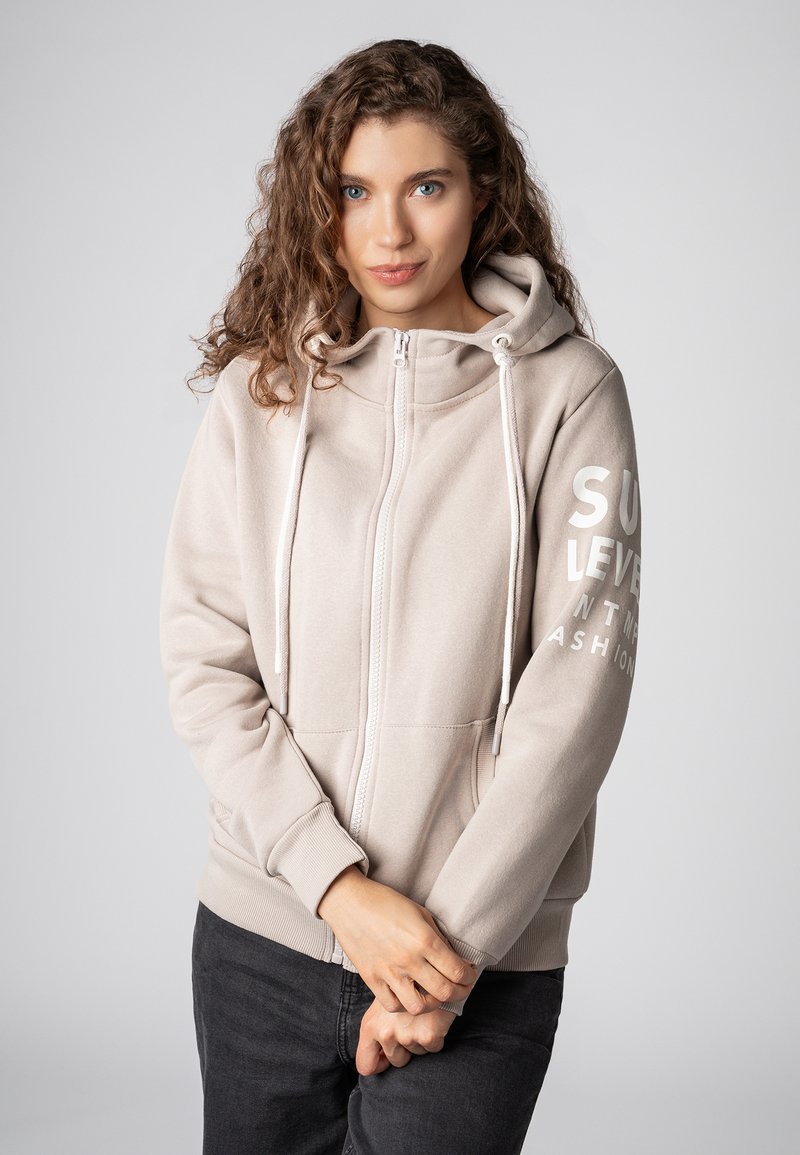 Lichtbeige zip-up hoodie met een trekkoordcapuchon, zachte textuur, frontzakken en witte bedrukte tekst op de linker mouw.
