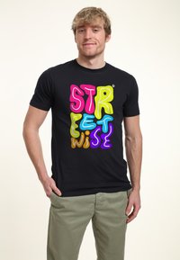 Camiseta de algodón negra con texto en estilo burbuja de múltiples colores que dice "STREETWISE". El portador la combina con pantalones de color claro.