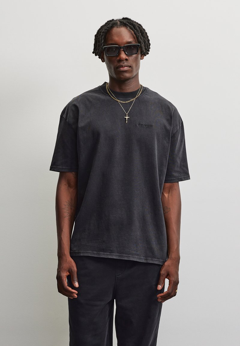 Mand med flettet hår iført sorte solbriller, en sort oversized t-shirt, sorte bukser og to gyldne halskæder med et korsvedhæng.