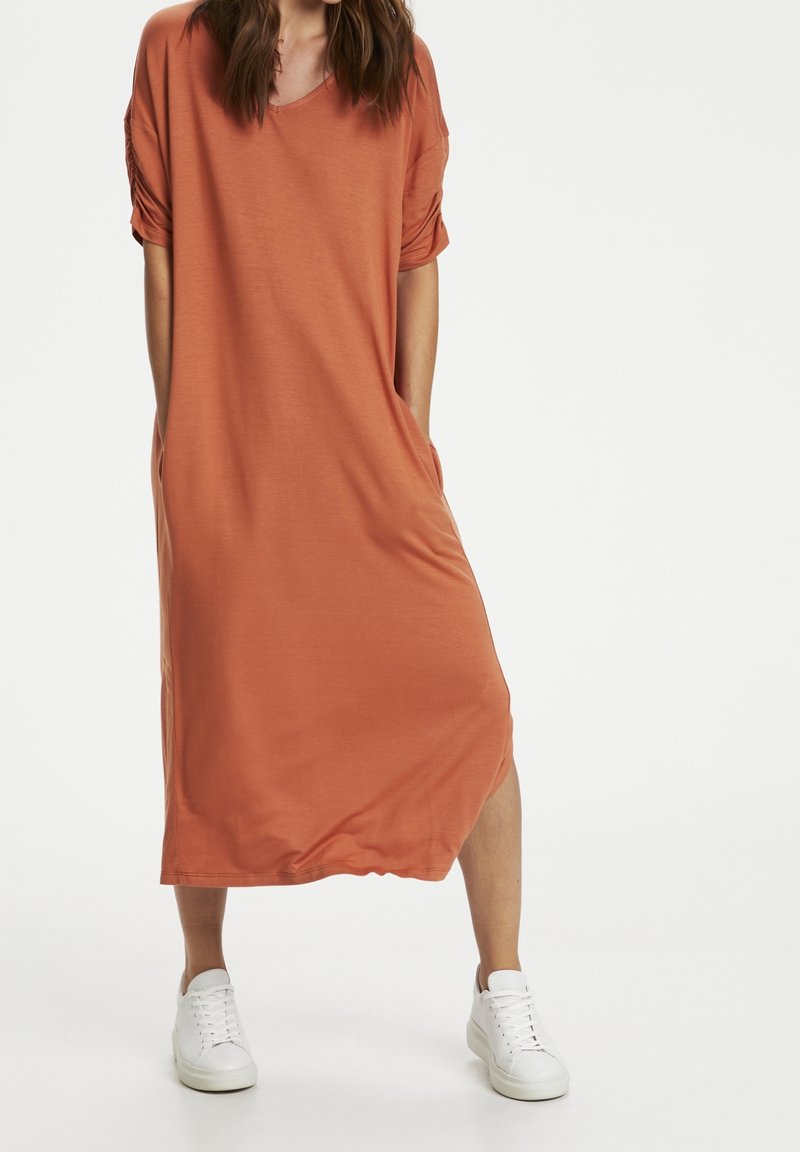 Femme portant une robe terracotta ample arrivant aux genoux avec des manches courtes et des baskets blanches, debout les mains dans les poches.