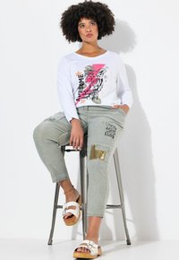 Haut à manches longues blanc avec graphique de tigre et texte, associé à un pantalon vert avec divers écussons et un accent doré. Sandales blanches aux pieds.