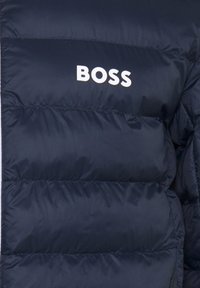 Section de veste matelassée bleu foncé avec des coutures horizontales et le mot BOSS en lettres blanches grasses sur le tissu.