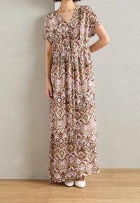 Robe maxi avec décolleté en V, manches courtes et taille élastique. Tissu à motifs en rose, vert et marron avec des designs géométriques. Chaussures blanches.