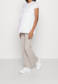 Hvid kortærmet graviditetstrøje med rund halsudskæring og knapdetalje, parret med lysebeige vide bukser og hvide sneakers.