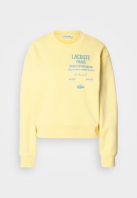 Sweat-shirt jaune Lacoste avec texte bleu incluant « LACOSTE PARIS FRENCH SPORTSWEAR » et un petit logo crocodile à l'avant.