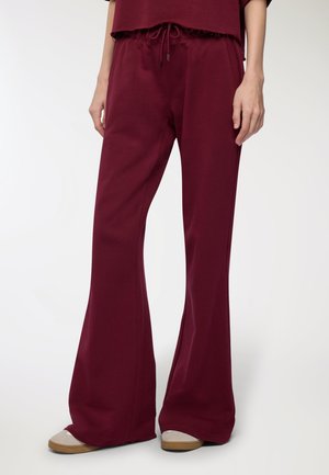 Pantaloni sportivi a zampa d'elefante color bordeaux realizzati in morbido cotone, dotati di vita con cordoncino e texture liscia, abbinati a scarpe beige senza lacci.
