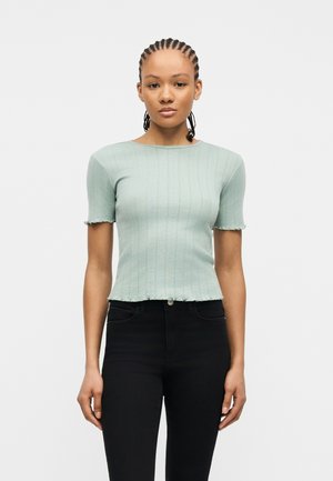 Mlada žena s pletenicama, odjevena u svijetlozelenu rebrastu majicu kratkih rukava i crne hlače visokog struka na jednobojnoj pozadini.