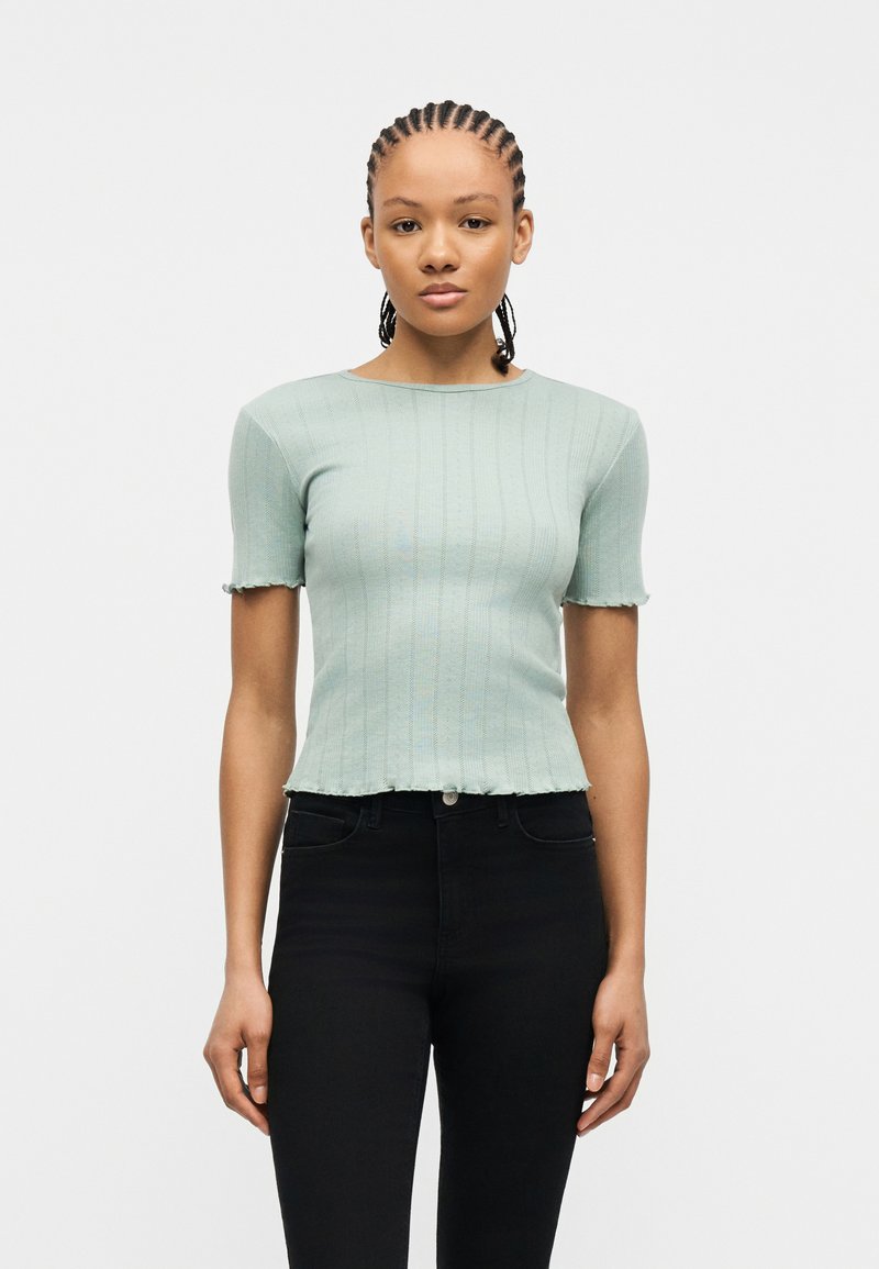 Νεαρή γυναίκα με πλεξούδες φοράει ένα ανοιχτοπράσινο ριμπ τοπ με κοντά μανίκια και ψηλόμεσο μαύρο παντελόνι, με φόντο ένα απλό φόντο.