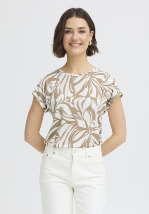 Smiling woman with short brown hair wears beige and white patterned short-sleeve top and high-waisted white pants, standing against white background.