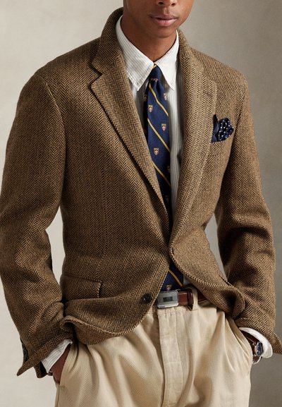 Brun blazer i fiskebensmønster med hakkede slag, kombinert med en stripete hvit skjorte, marineblå slips med mønster og beige bukser.