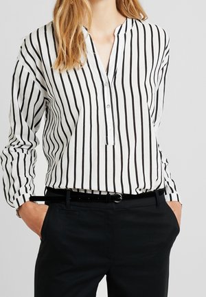 Kvinde iført en hvid bluse med lodrette sorte striber, sorte bukser og et tyndt sort bælte, hænderne i lommerne.