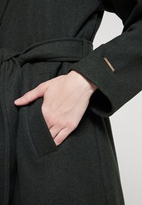 Abrigo de lana verde oscuro con cintura ceñida, bolsillos laterales y un diseño de puños elegante con un acento metálico cerca de la manga.