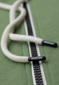 Gros plan sur des cordons blancs à embouts noirs portant l'inscription "DITABRE", reposant sur une fermeture éclair en tissu vert bordée de blanc.