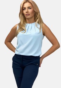 Jasnoniebieska bezrękawnik z plisowanym dekoltem, zestawiona z ciemnogranatowymi dopasowanymi spodniami z małymi złotymi akcentami.