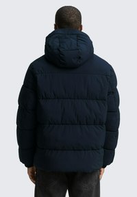 Giacca imbottita blu navy con cappuccio. Presenta sezioni trapuntate e polsini a costine. La vista posteriore mostra una silhouette squadrata e dettagli minimi.