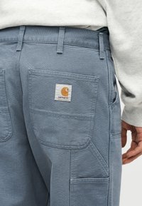 Άτομο που φοράει ανοιχτό μπλε παντελόνι Carhartt με πολλαπλές τσέπες και εμφανές λογότυπο μάρκας στην πίσω τσέπη, συνδυασμένο με ανοιχτόχρωμο πάνω μέρος.