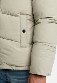Jasno beżowa kurtka puffer z pikowanym wzorem, z czarnym napem na kieszeni naszywanej oraz miękką, gładką teksturą.
