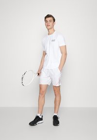 Balts sporta T-krekls ar EA7 logotipu, apvienots ar baltiem īsiem šortiem ar oranžiem akcentiem un melnām tenisa kurpēm; tur roka tenisa raketi.
