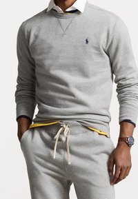 Hombre vestido con sudadera gris claro y pantalones deportivos a juego con cordón blanco, camiseta interior amarilla, cuello blanco y reloj de pulsera marrón.