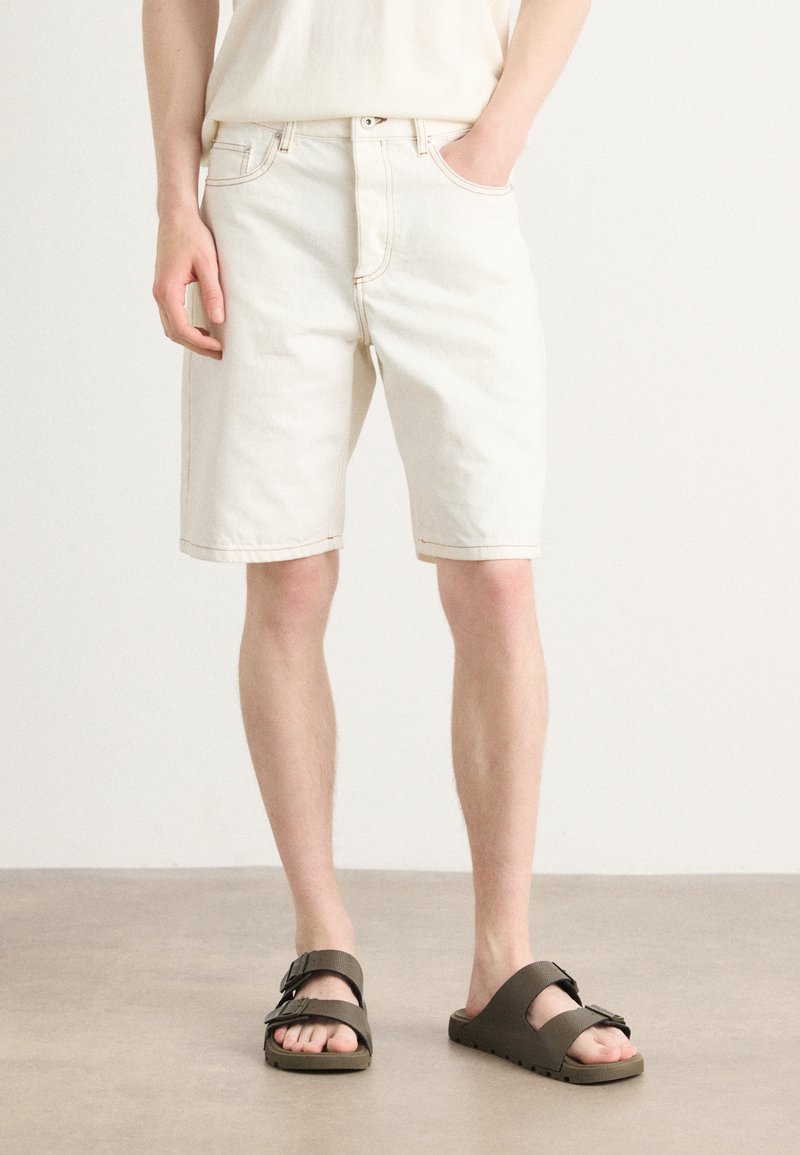 Shorts en denim blanc à coupe droite, dotés d'une fermeture par bouton et zip à l'avant, accompagnés de sandales marron à doubles brides.