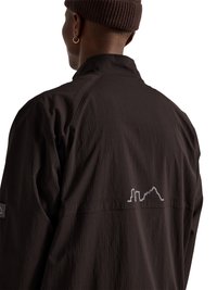 Mand iført en mørkebrun jakke og matchende strikhue, viser ryggen med et lille reflekterende bylandskabsdesign nær skulderbladene.