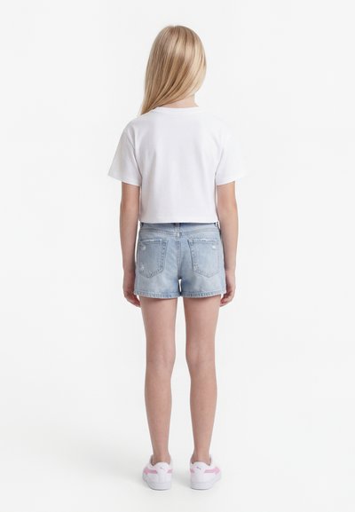 Niño con cabello largo y rubio de espaldas, vistiendo una camiseta blanca de manga corta lisa, shorts de mezclilla claros y zapatillas blancas con detalles en rosa.