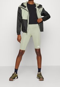 Jasnozielony wiatrówka z czarnymi akcentami, czarny t-shirt, jasnozielone szorty rowerowe i wielokolorowe buty sportowe. Widoczne teksturowane materiały.