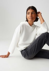 Vrouw die op de grond zit, gekleed in een witte textuur hoodie met driekwartmouwen en donkergrijze jeans, die zijwaarts kijkt met haar hand dicht bij haar oor.