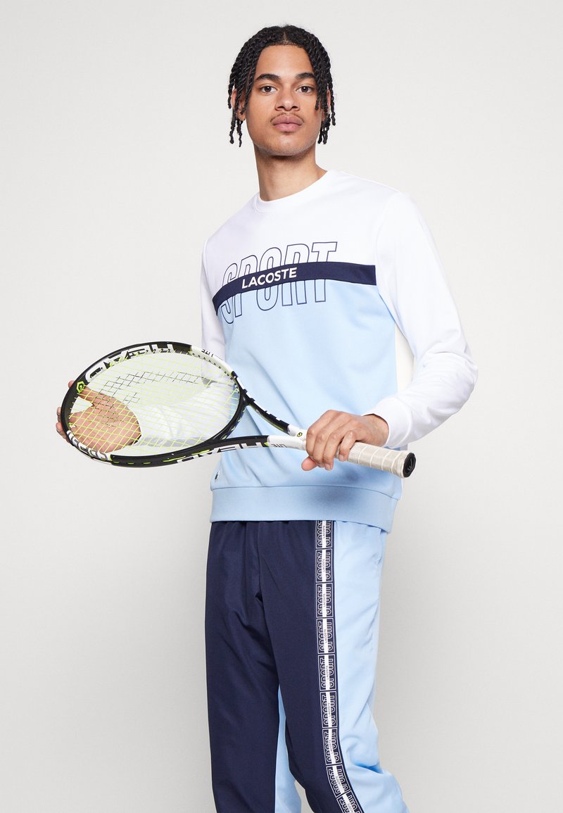 Conjunto deportivo en azul claro y marino, que incluye una sudadera con el texto "SPORT" y una raqueta de tenis sostenida en la mano derecha.