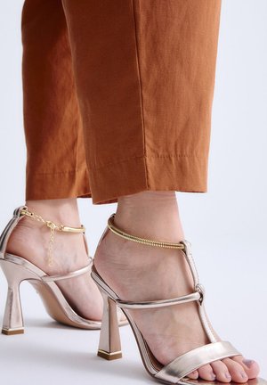Pies con sandalias metálicas de tacón alto y cadenas doradas en el tobillo, combinados con pantalones cortos de color óxido sobre un fondo liso.