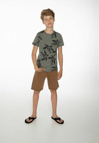 Grijs T-shirt met korte mouwen en zwart bladenpatroon, gecombineerd met bruine shorts en zwarte teenslippers. Model poseert met één hand in de zak.