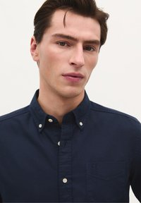 Navyblaues Button-up-Hemd aus Baumwolle mit Kragen, einer frontalen Tasche mit Logo und einer dezenten Textur. Schlichte Hintergrund.