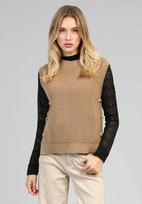 Pull en maille beige avec des manches longues en dentelle noire, comportant des boutons ronds sur le côté. Le design est ajusté avec une texture côtelée.