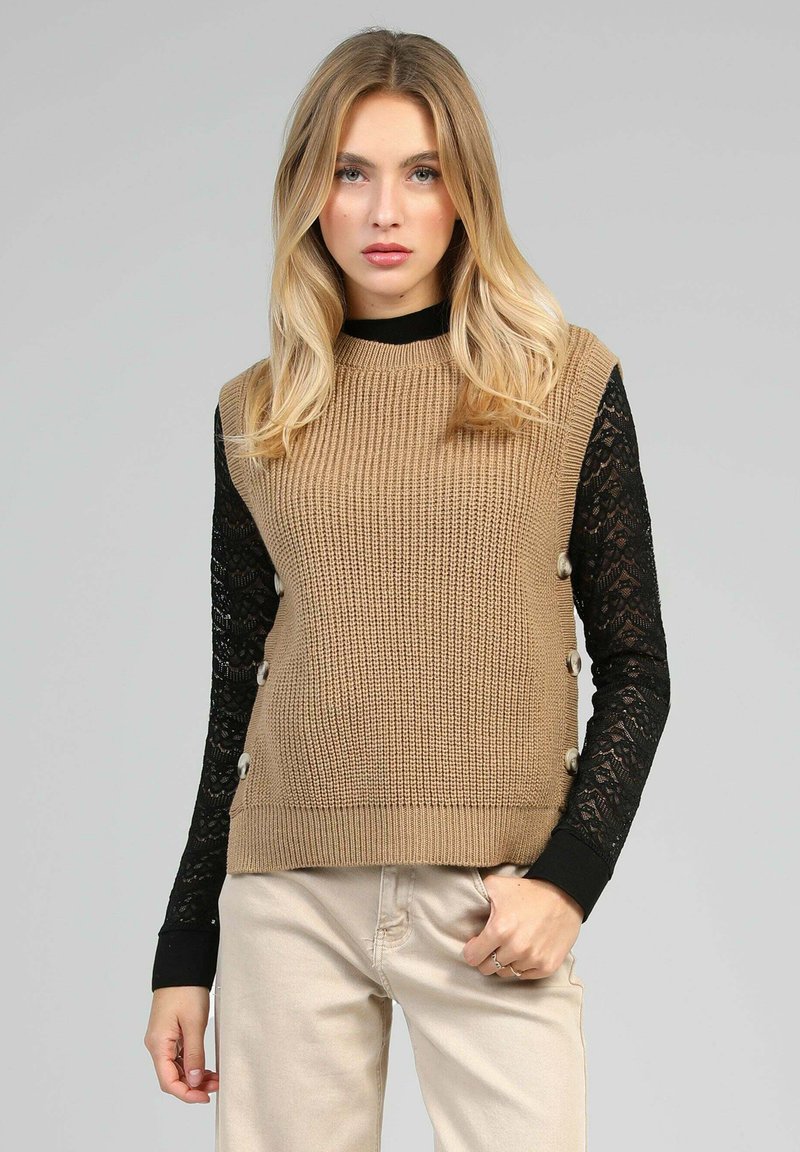 Pull en maille beige avec des manches longues en dentelle noire, comportant des boutons ronds sur le côté. Le design est ajusté avec une texture côtelée.