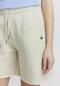 Beige joggeshorts laget av myk, teksturert stoff. Har elastisk midje med snor, sidelommer og en metallaccentdetalj.